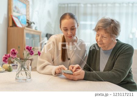 Young woman helping an senior woman with a smartphone at home. Concept of technology, education, and senior digital adaptation. Granddaughter teaches grandmother how to use digital device. Young woman helping an senior woman with a smartphone at home. Concept of technology, education, and senior digital adaptation. Granddaughter teaches grandmother how to use digital device. 123117211