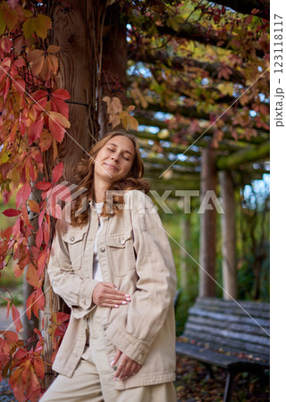 Autumn Portrait: Smiling Young Woman in Beige Outfit Leaning on Wooden Pergola with Red Leaves. Cozy Fall Scene Showcasing Natural Garden Background and Warm Seasonal Atmosphere in Rustic Outdoor Autumn Portrait: Smiling Young Woman in Beige Outfit Leaning on Wooden Pergola with Red Leaves. Cozy Fall Scene Showcasing Natural Garden Background and Warm Seasonal Atmosphere in Rustic Outdoor 123118117
