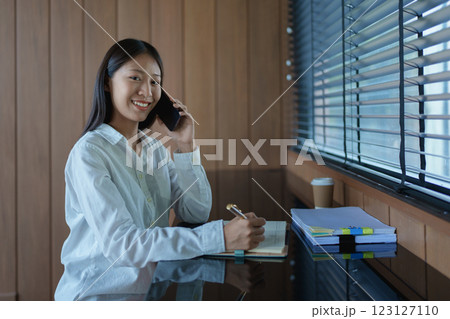 Young woman in casual white shirt smiling while talking on smartphone, working at desk with notepad and coffee 123127110