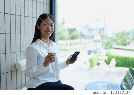 Young woman enjoying coffee while using smartphone in modern urban caf with laptop and natural light 123127124