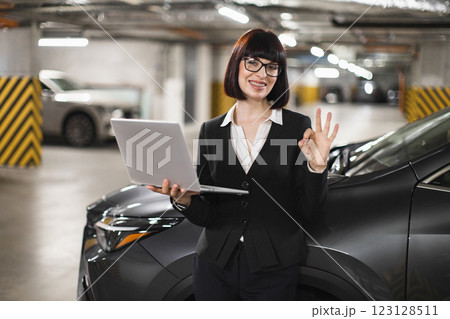Caucasian woman in professional attire stands in parking lot holding laptop, expressing confidence with OK gesture. Portrays modern professional workplace and technology in everyday business setting. Caucasian woman in professional attire stands in parking lot holding laptop, expressing confidence with OK gesture. Portrays modern professional workplace and technology in everyday business setting. 123128511