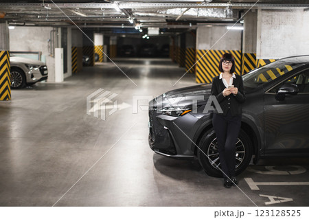 Confident young businesswoman using smartphone in modern car park. Caucasian female in business attire texting while standing next to car. Urban work life and connectivity. Confident young businesswoman using smartphone in modern car park. Caucasian female in business attire texting while standing next to car. Urban work life and connectivity. 123128525