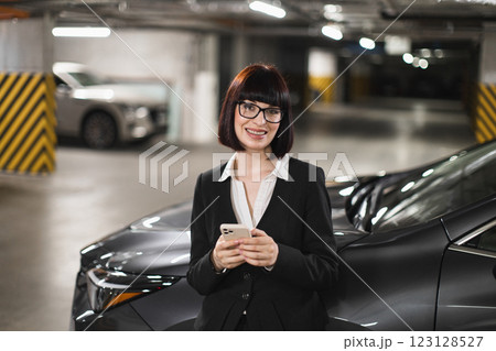 Confident young Caucasian business woman in suit standing in indoor car park, typing on phone, smiling confidently, showcasing professionalism and modern technology use. Confident young Caucasian business woman in suit standing in indoor car park, typing on phone, smiling confidently, showcasing professionalism and modern technology use. 123128527