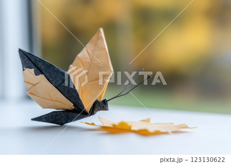 A detailed paper butterfly, folded from brown and black sheets, sits gracefully on a white surface. The background features soft, blurred autumn colors, enhancing the tranquility of the moment 123130622