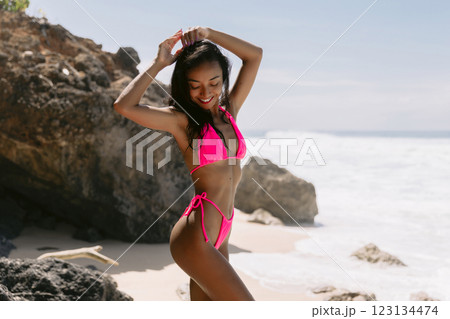 Smiling woman in a bikini enjoying a sunny beach day, embracing summer vibes with ocean waves and rocky cliffs behind her Smiling woman in a bikini enjoying a sunny beach day, embracing summer vibes with ocean waves and rocky cliffs behind her 123134474