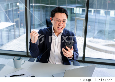 Excited Asian Businessman Celebrating Success Using Smartphone in Modern Office 123134727