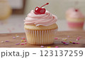 Golden vanilla cupcake with soft pink frosting, topped with a glossy red cherry and colorful sprinkles, placed on rustic wooden surface with scattered sprinkles, blurred background with more cupcakes, 123137259