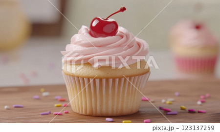 Golden vanilla cupcake with soft pink frosting, topped with a glossy red cherry and colorful sprinkles, placed on rustic wooden surface with scattered sprinkles, blurred background with more cupcakes, 123137259