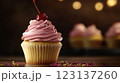 Vanilla cupcake with deep pink frosting and glossy red cherry, moody lighting enhancing golden cake crumb, placed on dark textured wooden backdrop with scattered colorful sprinkles, blurred background 123137260