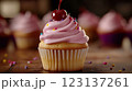 Vanilla cupcake with deep pink frosting and glossy red cherry, moody lighting enhancing golden cake crumb, placed on dark textured wooden backdrop with scattered colorful sprinkles, blurred background 123137261