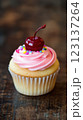 Vanilla cupcake with deep pink frosting and glossy red cherry, moody lighting enhancing golden cake crumb, placed on dark textured wooden backdrop with scattered colorful sprinkles, blurred background 123137264