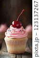 Vanilla cupcake with deep pink frosting and glossy red cherry, moody lighting enhancing golden cake crumb, placed on dark textured wooden backdrop with scattered colorful sprinkles, blurred background 123137267