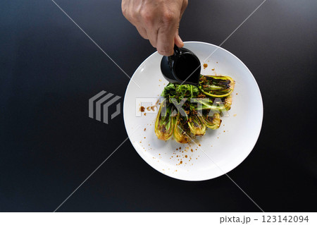 A hand pours sauce over fresh bok choy arranged on a plate, highlighting Asian cooking techniques. A hand pours sauce over fresh bok choy arranged on a plate, highlighting Asian cooking techniques. 123142094