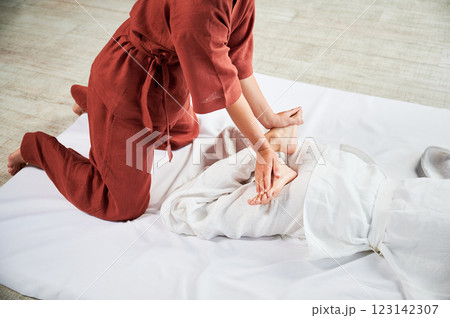 Masseuse making thai yoga massage. Woman therapist doing traditional massage treatment, stretching female patient's body. Client lying on mat, enjoying hand massage in calm atmosphere. Cropped view. 123142307
