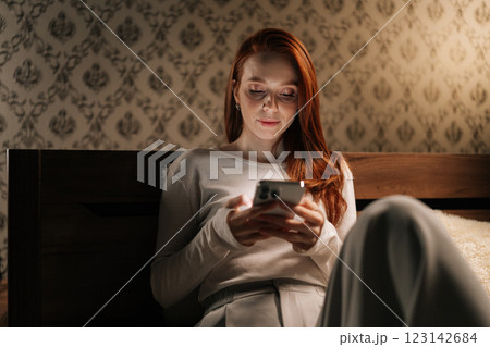 Focused tired young woman using browsing smartphone sitting on bed in dark bedroom at night, light from screen illuminating face. Concept of social media addiction, phone overuse, insomnia. Focused tired young woman using browsing smartphone sitting on bed in dark bedroom at night, light from screen illuminating face. Concept of social media addiction, phone overuse, insomnia. 123142684