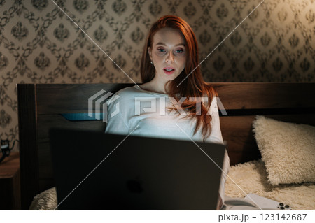 Redhead young woman looking surprised while reading shocking news on laptop, sitting in bed at night, illuminated by screen light, experiencing moment of astonishment and disbelief. Redhead young woman looking surprised while reading shocking news on laptop, sitting in bed at night, illuminated by screen light, experiencing moment of astonishment and disbelief. 123142687