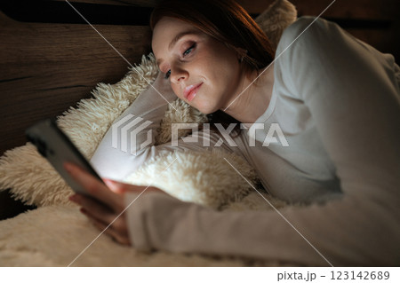 Peaceful redhead woman lying on bed, using smartphone in dark bedroom at night, enjoying leisure time while smiling looking to illuminated screen, radiating happiness. Peaceful redhead woman lying on bed, using smartphone in dark bedroom at night, enjoying leisure time while smiling looking to illuminated screen, radiating happiness. 123142689