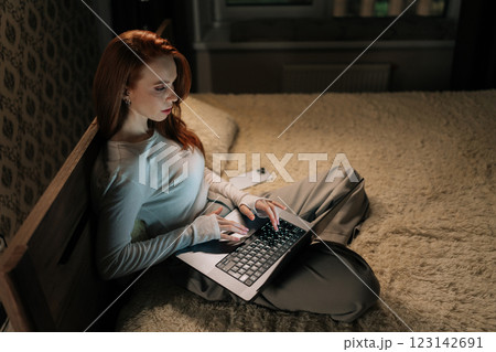 Top view of focused female freelancer working late on laptop sitting in bed, face illuminated by screen glow in bedroom, demonstrating dedication to tasks. Successful woman discussing new project 123142691