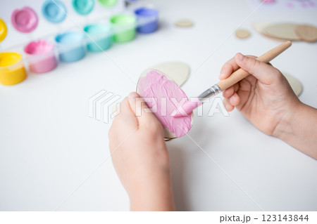Child painting Easter wooden egg with pink brush on white table with colorful paints 123143844