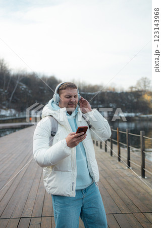 Smiling young man is walking near a lake on the forest, wearing headphones and using a mobile phone. Smiling young man is walking near a lake on the forest, wearing headphones and using a mobile phone. 123143968