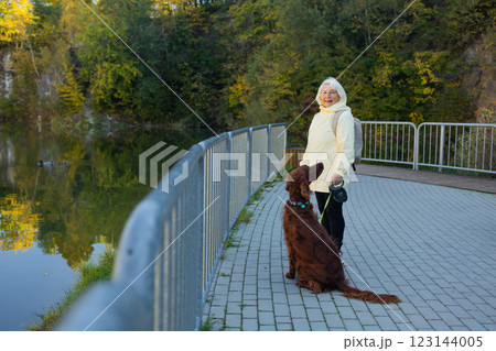 Happy senior 60s woman crossbreed looking out over a lake with her best friend irish setter dog wear casual clothes in the autumn forest.  123144005