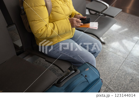 A cropped image of a beautiful Caucasian female tourist is holding her smartphone and passport, checking her flight information on her phone while sitting at a waiting seat in the airport terminal. 123144014