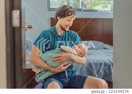 A father sits on a fitness ball in a cozy bedroom, gently rocking his baby wrapped in a soft muslin swaddle. A nurturing and soothing moment of fatherhood and care. Parenting, bonding, and comfort 123144574