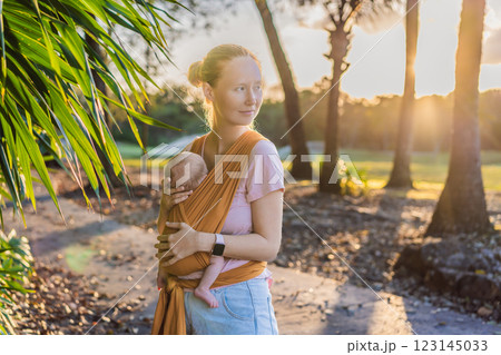 A loving mother walks through a peaceful park, carrying her baby securely in a sling. A heartwarming moment of maternal bonding, babywearing, and the joy of motherhood in nature 123145033