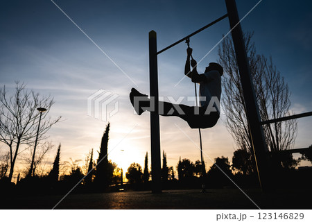 young athletic man exercising fitness calisthenics workout and making ring leg raises for abdomen abs muscles at street gym during sunset, body improvement and strength training 123146829