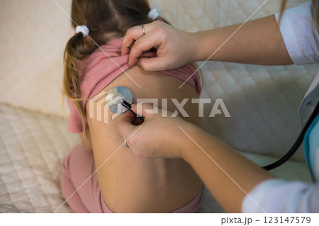 A female pediatrician uses a stethoscope to examine the lungs of a sick little girl sitting on a bed during a home visit. The pediatrician provides personalized medical care to a sick child at home. A female pediatrician uses a stethoscope to examine the lungs of a sick little girl sitting on a bed during a home visit. The pediatrician provides personalized medical care to a sick child at home. 123147579