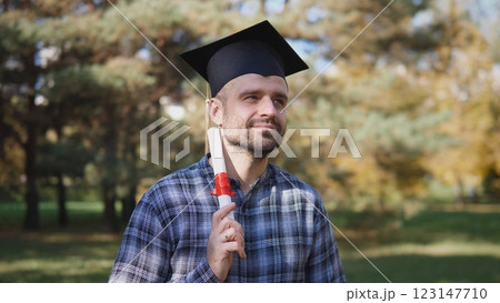 A joyful man dressed in graduation attire is holding his diploma proudly outdoors amidst the vibrant autumn foliage A joyful man dressed in graduation attire is holding his diploma proudly outdoors amidst the vibrant autumn foliage 123147710