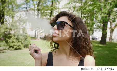 In a vibrant park, a stylish woman smokes leisurely, radiating a sense of confidence and relaxation effortlessly 123147743