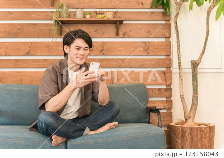 家のリビングでスマホを見ながらくつろぐ笑顔の若いアジア人・日本人男性（リラックス） 123158043