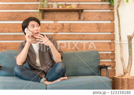 ソファでくつろぎながらスマホで通話する若いアジア人男性（驚く・びっくり・衝撃・バレる・怒られる） 123158180