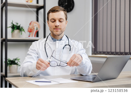 A young attractive doctor sits at his workplace in the office and takes off his glasses at the end of the working day 123158536