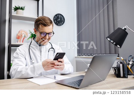 Male doctor medical worker in modern clinic wearing eyeglasses and white coat uniform using cell mobile smartphone apps, sitting at laptop computer. Medicine technologies health care concept. 123158558