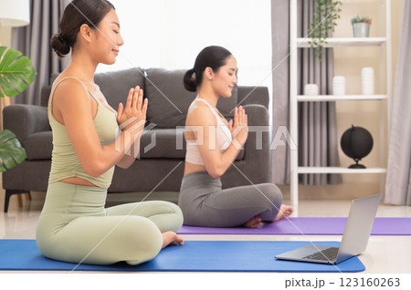 Two women doing yoga meditating exercise in living room, sitting in Easy seat pose with mudra in  bedroom. Working out wearing sportive top and leggings with laptop on blue mat. Relaxation and 123160263