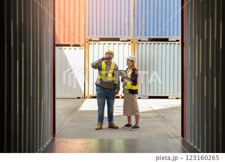 Two workers wearing safety vests and hard hats while standing in shipping container yard. Male holding walkie talkie and female using digital tablet for cargo loading information 123160265