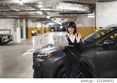 Caucasian woman in smart attire using laptop for video call in indoor parking lot. Professional setting with vehicles in background, reflecting modern business environment. 123160631