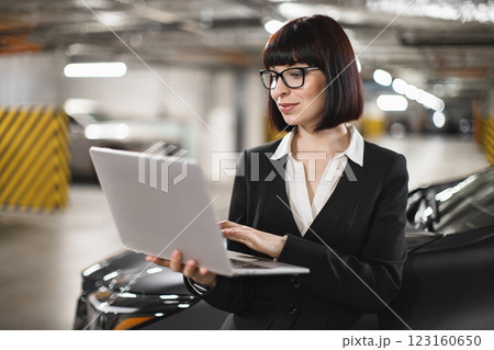 Caucasian businesswoman in her 30s using laptop in indoor parking lot, focused and professional. She wears glasses and business attire, standing beside car. Concept of mobile work and technology. Caucasian businesswoman in her 30s using laptop in indoor parking lot, focused and professional. She wears glasses and business attire, standing beside car. Concept of mobile work and technology. 123160650