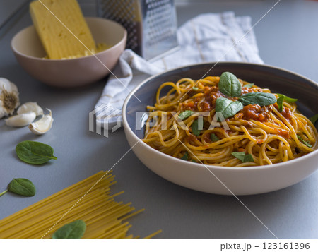 Balanesi pasta nestled in contemporary ceramic bowl, adorned with basil leaves, surrounding raw spaghetti, garlic, spinach, grated cheese on minimalist grey surface Balanesi pasta nestled in contemporary ceramic bowl, adorned with basil leaves, surrounding raw spaghetti, garlic, spinach, grated cheese on minimalist grey surface 123161396