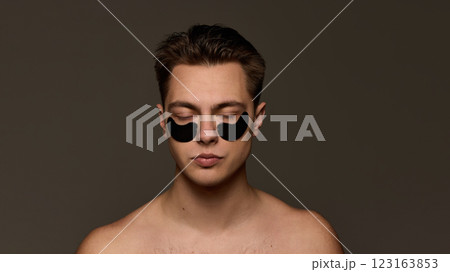 Young man with closed eyes, with black hydrogel eye patches, radiating relaxation and rejuvenation in skincare against brown studio background. Young man with closed eyes, with black hydrogel eye patches, radiating relaxation and rejuvenation in skincare against brown studio background. 123163853