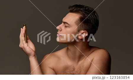 Serious young man holding gold spraying bottle near forehead, exuding luxury and grooming precision, against brown studio background. 123163854