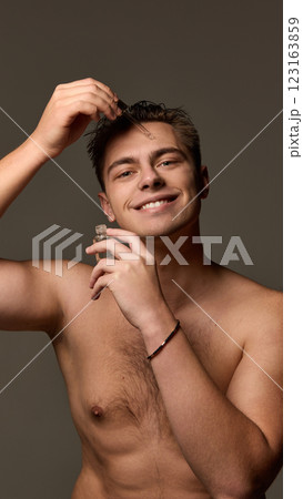 Man smiling while applying serum with dropper, holding bottle in other hand, looking into camera against brown studio background. Man smiling while applying serum with dropper, holding bottle in other hand, looking into camera against brown studio background. 123163859