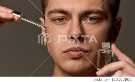 Close-up of man applying serum under eye with dropper, liquid running down cheek, focused expression against brown studio background. Close-up of man applying serum under eye with dropper, liquid running down cheek, focused expression against brown studio background. 123163860