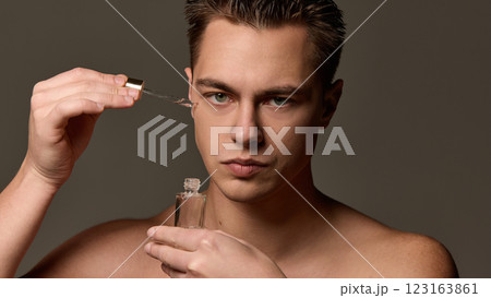 Shirtless man with serious expression applying serum with dropper near eye, looking directly at camera against brown studio background. 123163861