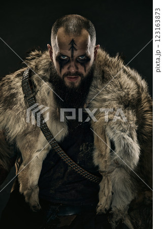 Close-up portrait of Viking warrior with war paint, piercing blue eyes, and fur cloak, staring menacingly against dark background 123163873