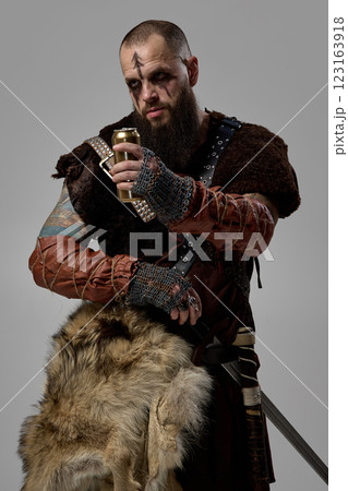 Portrait of Viking warrior gripping golden beer bottle, wearing fur cloak and battle-worn leather armor against studio background. Portrait of Viking warrior gripping golden beer bottle, wearing fur cloak and battle-worn leather armor against studio background. 123163918
