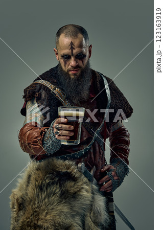 Portrait of medieval Viking warrior dressed in fur cloak and leather armor, standing with sword, holding glass of beer, against studio background. Portrait of medieval Viking warrior dressed in fur cloak and leather armor, standing with sword, holding glass of beer, against studio background. 123163919