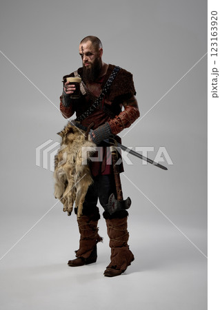Viking warrior standing with sword and axe, holding glass filled with beer, dressed in fur cloak and leather armor against studio background. 123163920
